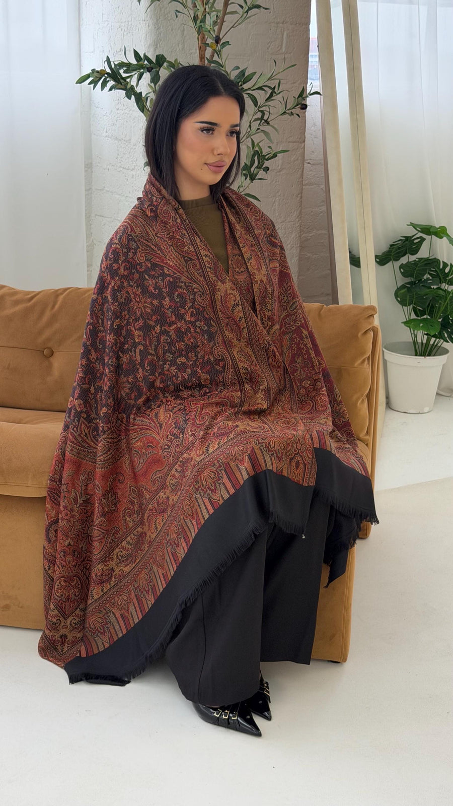 Woman sitting on a couch wearing a black pashmina patterned shawl in a home setting