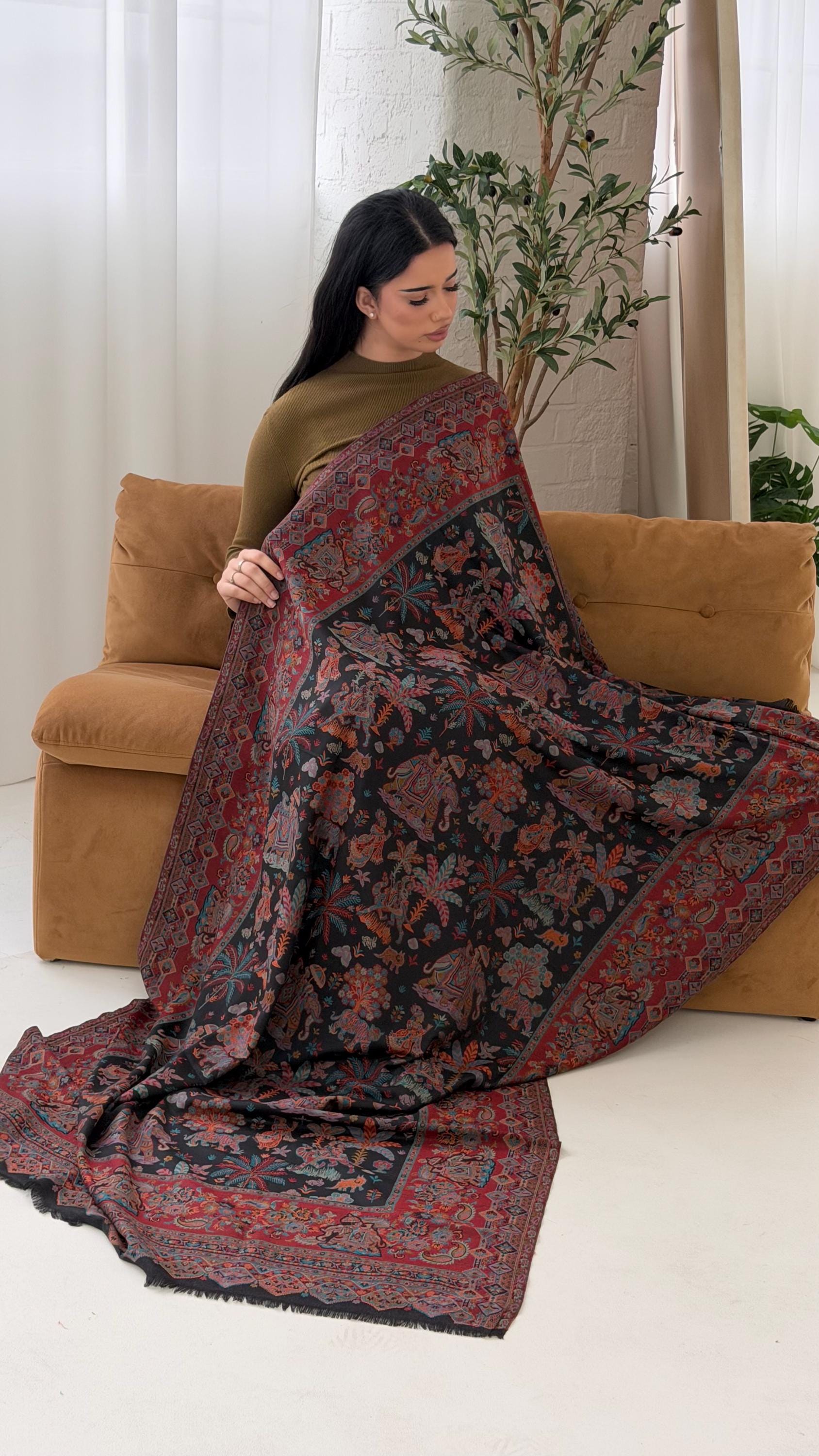 Woman holding a black patterned pashmina shawl scarf in a room with a couch and plants.